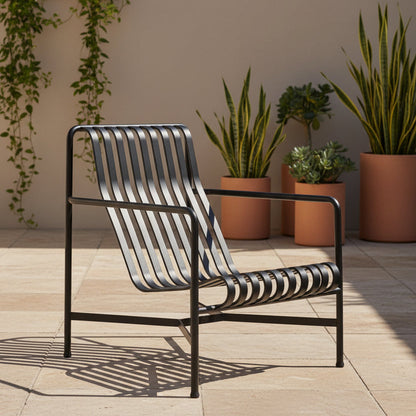 A modern outdoor lounge chair with a black metal frame and woven slatted seat and back, positioned on a light stone patio in front of a beige wall with potted plants.